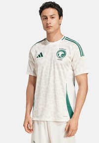 Maglia sportiva bianca con una sottile stampa leopardata, dettagli verdi e righe. Presenta un collo rotondo e il logo "Saudi" sul petto.