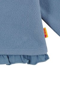 Fleece'owa bluzka w miękkim niebieskim kolorze z fakturowanej tkaniny, z falbanką u dołu w dopasowanym kolorze oraz małą, żółtą metką z boku.