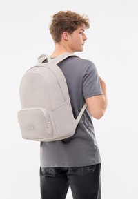 Szürke hátizsák sima, matt felülettel, kerek formával, elülső cipzáras zsebbel és állítható párnázott pántokkal. Szürke felsőruházat felett viselve.