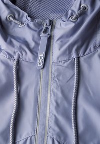 Veste violet clair avec un devant zippé, des cordons de serrage et une doublure en mesh. Présente un tirant de logo et des accents réfléchissants sur la fermeture éclair.