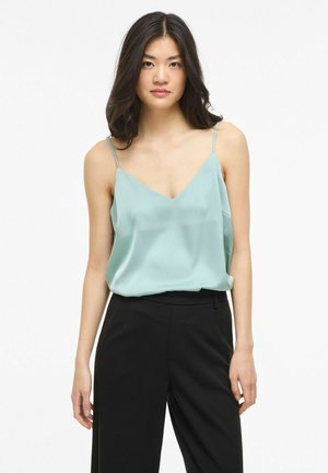 Jeune femme aux longs cheveux foncés portant un caraco en satin bleu clair et un pantalon noir taille haute, debout devant un fond blanc.