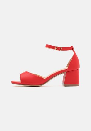 Rød åben sandal med chunky mellemhæl og justerbar ankelrem med guldlås på hvid baggrund.