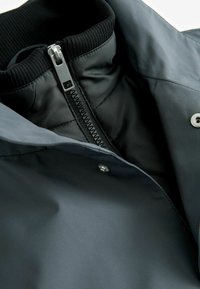 Veste grise avec un col côtelé noir, dotée d'une fermeture éclair métallique et de détails à bouton-pression. Tissu lisse avec un léger éclat subtil.