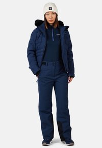 Marinblå isolerad jacka med foder av päls i huvan, i kombination med matchande marinblå byxor med dragkedja och knappstängning. Beigefärgad stickad mössa på huvudet.