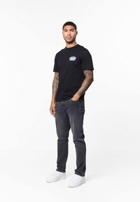 Svart t-shirt med en liten grafisk tryckning på vänster bröst, kombinerad med mörkgrå jeans och vita sneakers. Modellen står i en neutral miljö.