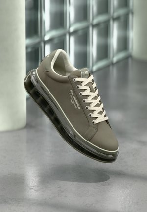 Zapatilla gris Karl Lagerfeld con cordones blancos y suela transparente amortiguada con aire flotando sobre un suelo de hormigón pulido.