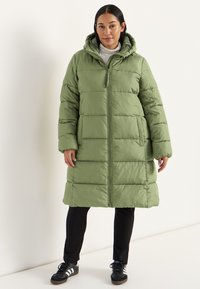 Kvinde iført en lang, olivengrønn quilted pufferjakke med hætte, sorte bukser, sort og hvid sneakers, der står mod en ensfarvet lys baggrund.