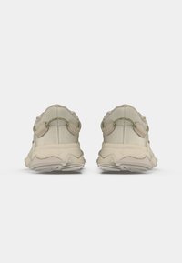 Un paio di sneakers beige casual viste da dietro, con colli imbottiti, suole spesse e testurizzate, e linguette su uno sfondo bianco.