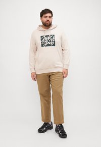 Beżowa bluza z kapturem z nadrukiem na środku, w zestawieniu z beżowymi spodniami khaki. Całość dopełniają czarno-białe buty sportowe.