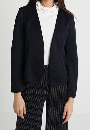 Femme portant un blazer noir ouvert sur le devant, un haut blanc à volants et col montant, et un pantalon noir à jambes larges avec de fines rayures verticales.