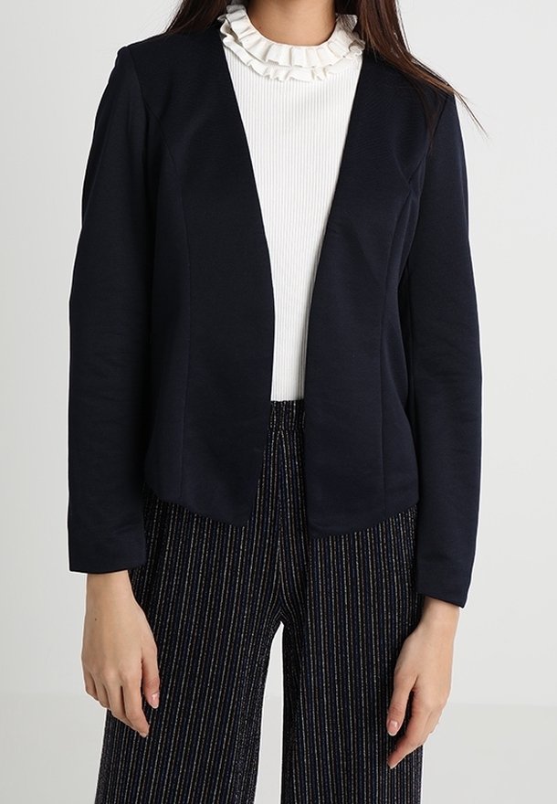 Femme portant un blazer noir ouvert sur le devant, un haut blanc à volants et col montant, et un pantalon noir à jambes larges avec de fines rayures verticales.