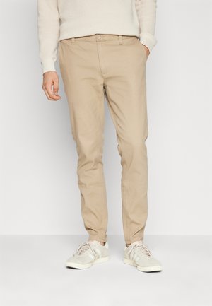 Beige Baumwollhosen mit slim fit, ausgestattet mit seitlichen Taschen und einem Knopfverschluss. Kombiniert mit hellen Sneakers.
