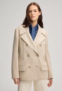 Beige dubbelzijdig colbert met grote revers, twee zijzakken en knoopaccenten. Gelaagd over een blauw denim top.