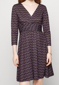 Klänning med V-ringning i mörklila tyg, med ett blommönster i grönt och guld. Långa ärmar och rynkad midja för en figurnära silhuett.