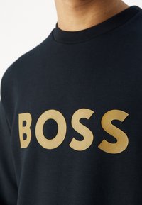 Acercamiento de una camisa azul marino con grandes y llamativas letras doradas que dicen "BOSS" en el pecho.