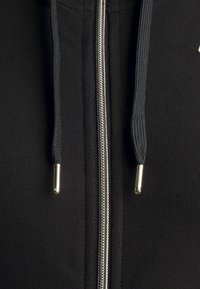 Svart zip-up hoodie med silverfärgad dragkedja, texturerat tyg och mörka snören med metalliska ändar på en enhetlig bakgrund.