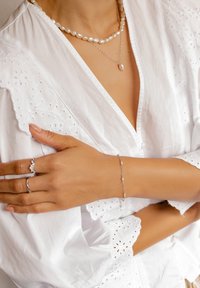 Blouse blanche avec des détails en broderie. Les bijoux comprennent un collier en perles, un bracelet en argent et deux bagues en argent avec des accents en diamant. Textures douces.