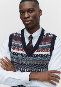 Stickad väst med ett mönster i blått, vinrött och krämfärgat, bärs över en ljusblå skjorta med krage och tunna ränder.