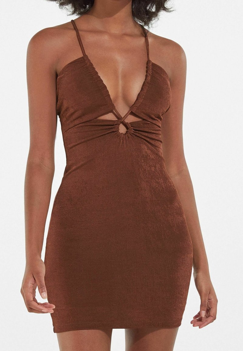 Femme portant une mini-robe marron sans manches avec de fines bretelles et une petite découpe à l'avant, sous la poitrine.