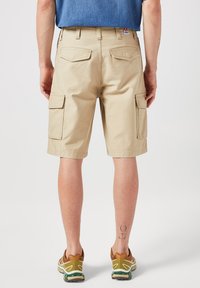 Pantaloni cargo beige con doppie tasche posteriori, orlo risvoltato e tasche laterali. Indossati con sneakers marroni con accenti verdi e un tatuaggio visibile.