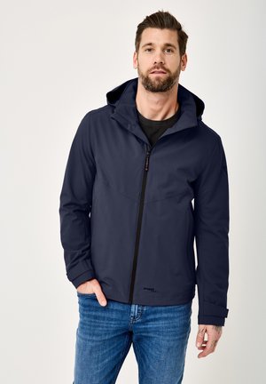 Mann mit Bart trägt eine marineblaue Kapuzenjacke und blaue Jeans, steht mit einer Hand in der Tasche vor einem schlichten Hintergrund.