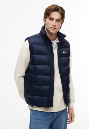 Ung mand iført en marineblå dynevest over en beige langærmet skjorte og blå jeans, står mod en hvid baggrund.