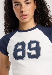 Camiseta blanca de manga corta raglán en azul marino con un gran gráfico "89" en azul marino contorneado en negro.