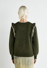 Personne aux cheveux blonds courts portant un pull vert foncé à épaules volantées et une jupe à rayures verticales de couleur claire, vue de dos.