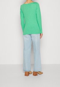 Maglia verde a maniche lunghe abbinata a jeans a gamba larga blu chiaro e sandali con tacco a blocco beige, mostrata da dietro.