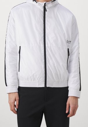 Homme portant une veste à capuche blanche zippée avec des bandes noires sur les manches et des accents de fermeture éclair noirs, associée à un pantalon noir.