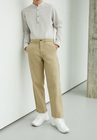 Beige bomullbyxor med rak skärning och sidfickor, kombinerade med en ljusgrå långärmad skjorta med knappstängd halsringning. Vita sneakers.