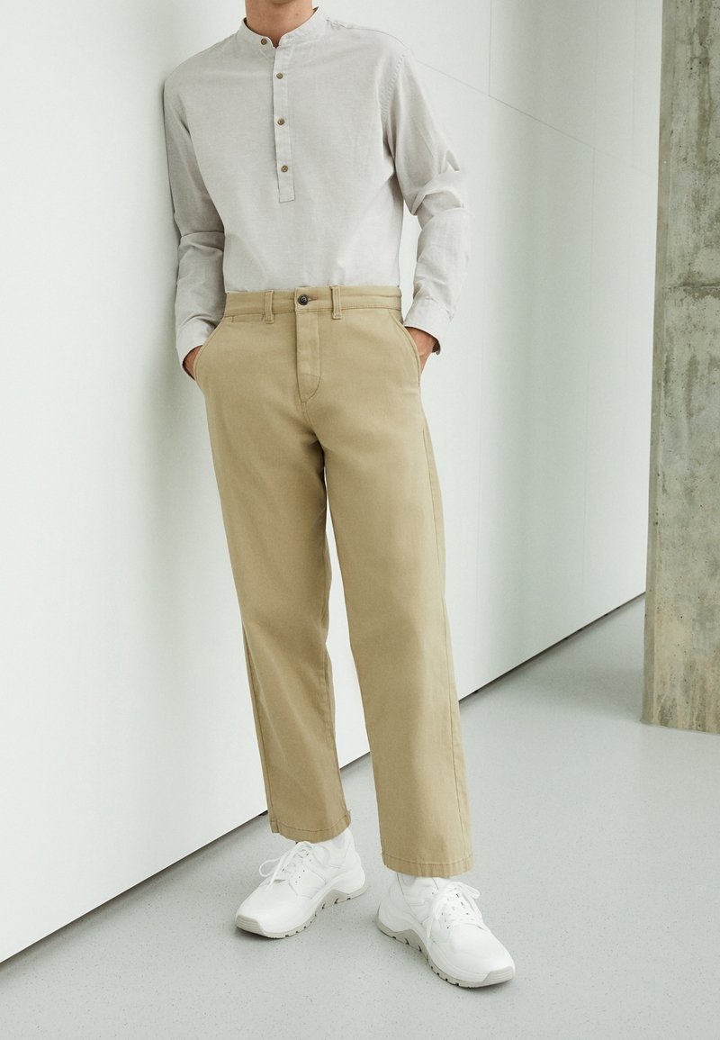 Beige bomullbyxor med rak skärning och sidfickor, kombinerade med en ljusgrå långärmad skjorta med knappstängd halsringning. Vita sneakers.