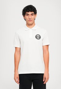 Νεαρός άνδρας με σκούρα σπυρωτά μαλλιά, φορώντας ένα άσπρο πόλο μπλουζάκι με ένα μαύρο κυκλικό λογότυπο Versace Jeans Couture στο στήθος, στέκεται μπροστά από ένα απλό φόντο.