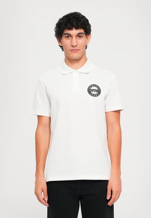 Tânăr cu păr creț închis la culoare, purtând un tricou polo alb cu un logo circular negru Versace Jeans Couture pe piept, stând pe un fundal simplu.