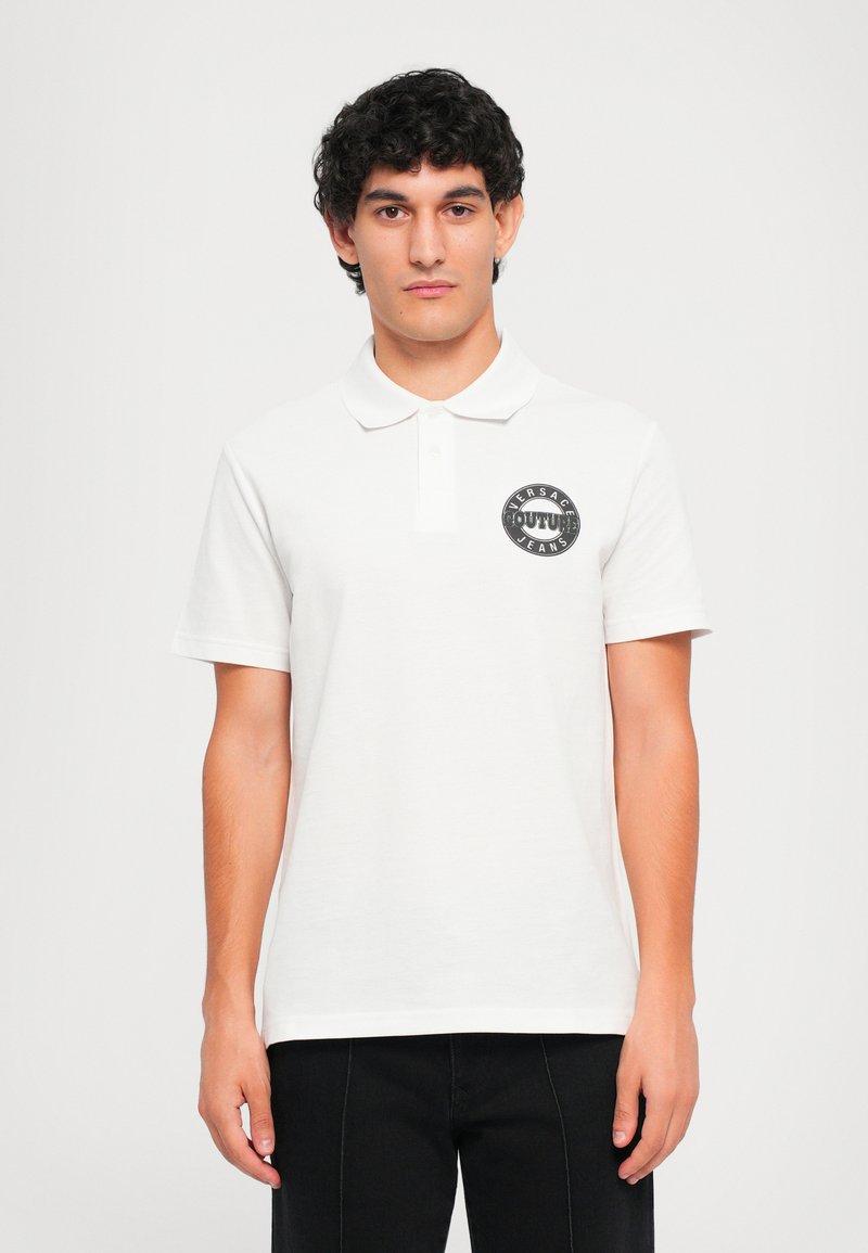 Νεαρός άνδρας με σκούρα σπυρωτά μαλλιά, φορώντας ένα άσπρο πόλο μπλουζάκι με ένα μαύρο κυκλικό λογότυπο Versace Jeans Couture στο στήθος, στέκεται μπροστά από ένα απλό φόντο.