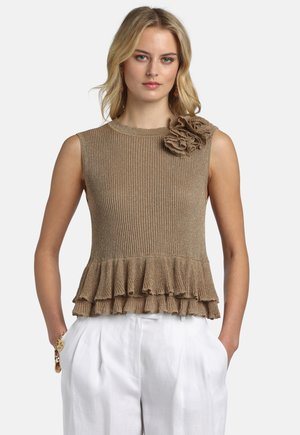 Lyshåret kvinde iført brun, ærmeløs ribstrikket top med flæsekant og blomsterdetalje, kombineret med hvide, højtaljede bukser.