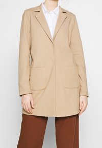 Beżowa marynarka z klapami, zapinana na jeden guzik, z dwoma naszytymi kieszeniami z przodu. Gładka tekstura i dopasowany krój.