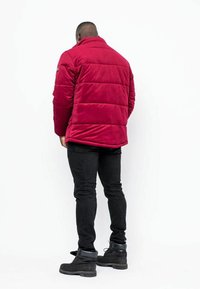 Giacca imbottita rossa con colletto alto, struttura sottile e cuciture visibili. Abbinata a jeans neri e stivali neri stringati.