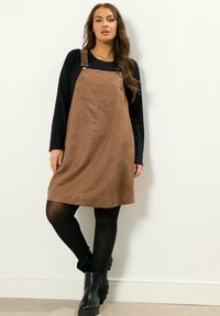 Un vestido de tirantes de ante marrón con tirantes ajustables, bolsillo frontal y corte en A, llevado sobre una blusa negra de manga larga y combinado con botas negras.