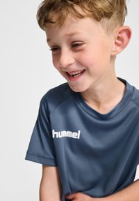 Szara sportowa koszulka wykonana z gładkiej tkaniny, z białym logo na lewej piersi i okrągłym dekoltem. Krótkie rękawy, luźny krój.