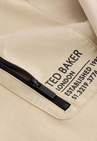Beżowy materiał o teksturowym wykończeniu, z czarnym zamkiem błyskawicznym i metką. Na metce widnieje napis "TED BAKER LONDON" oraz szczegóły dotyczące założenia firmy.