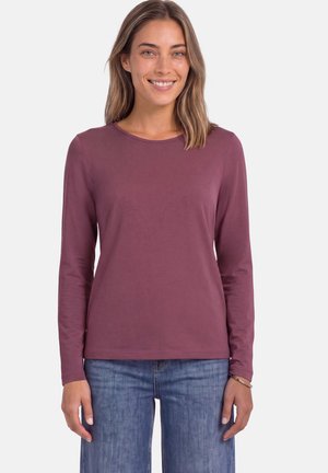 Femme aux cheveux châtain clair mi-longs, portant une chemise mauve à manches longues et un jean bleu, souriante devant un fond uni.