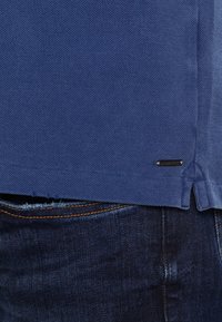 Blaues Hemd mit strukturiertem Finish, mit einem Saumdetail und einem kleinen schwarzen Logotag am unteren Ende. Kombiniert mit dunklen Jeans.