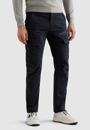 Uomo che indossa pantaloni cargo blu scuro con molte tasche e sneakers bianche, in piedi davanti a uno sfondo chiaro e uniforme.