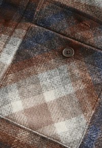 Braun-graues und blaues kariertes Gewebe mit einer weichen Textur. Enthält einen braunen Knopf und ausgeprägte Nähte. Verfügt über ein quadratisches Mustermuster.