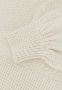 Maglione lavorato a maglia color crema con polsini a costine e un sottile dettaglio a palloncino sulle spalle, che mostra una texture morbida e un motivo minimale.