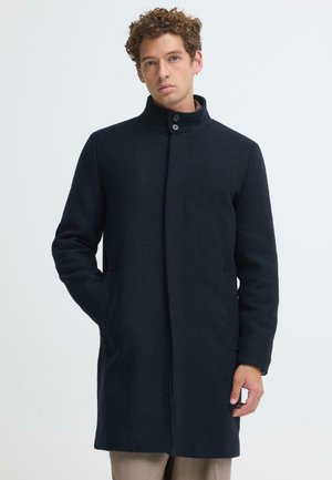 Homme aux cheveux bouclés portant un manteau bleu marine foncé boutonné avec un col montant et les mains partiellement dans les poches sur un fond uni.