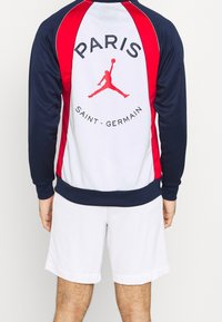 Trainingsjack met marineblauwe mouwen, witte romp met rode accenten, tekst "PARIS" en "SAINT-GERMAIN". Jumpman-logo in rood op de achterkant.