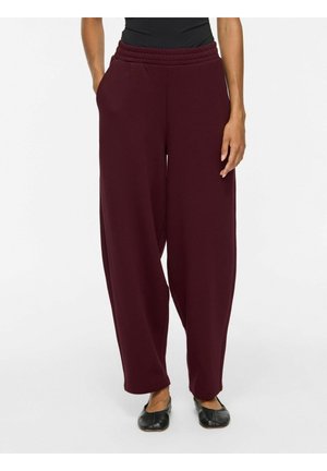Femme portant un pantalon large bordeaux avec une taille élastique et des chaussures plates noires, debout devant un fond blanc uni.