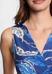 Robe bleue à motifs avec un décolleté en V, ornée de motifs de feuilles blanches. Un gros plan montre une texture lisse et un design sans manches.